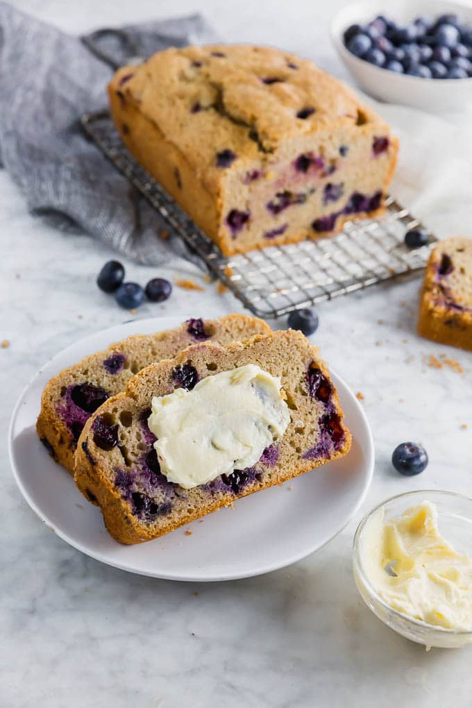 A loaf of blueberry muffin bread with two slices on a small plate slated in dairy free butter.