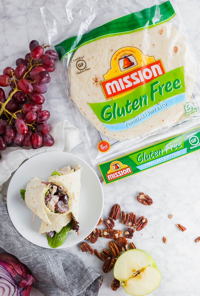A photo of two gluten free chicken salad wraps on a plate with a package of Mission Gluten Free tortillas and red grapes.