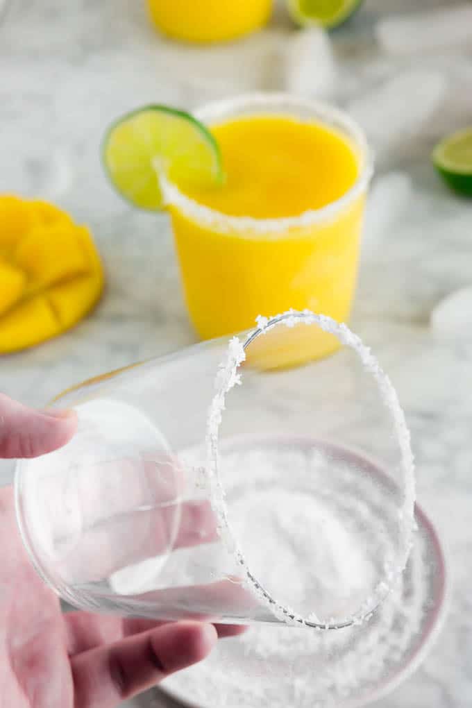 A photo of a margarita glass being rimmed with salt with a glass of frozen mango margarita in the background.