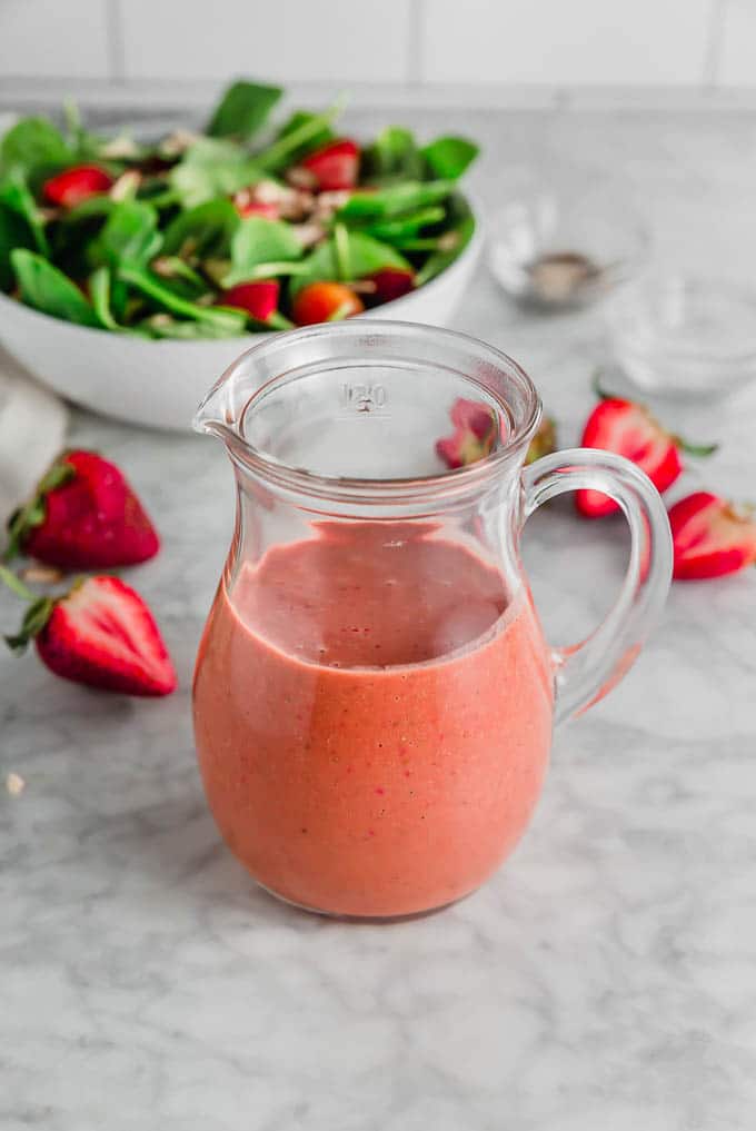 A photo of a glass pitcher with strawberry balsamic vinaigrette with fresh sliced strawberries and a spinach salad in the background.