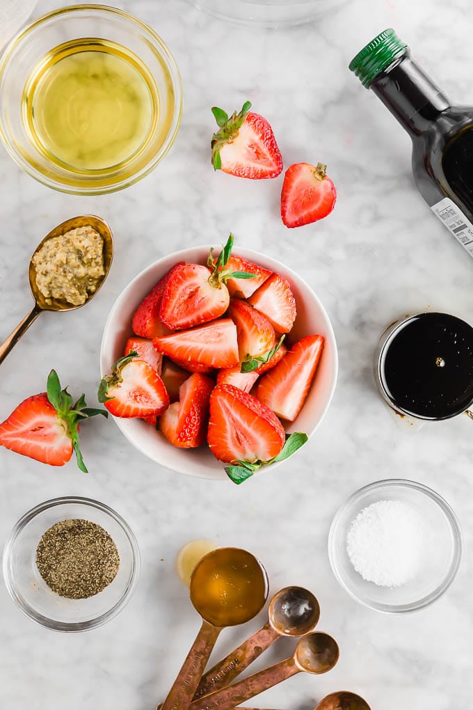 A photo of sliced strawberries, ground mustard, olive oil, balsamic vinegar, salt, pepper and honey on a marble table.