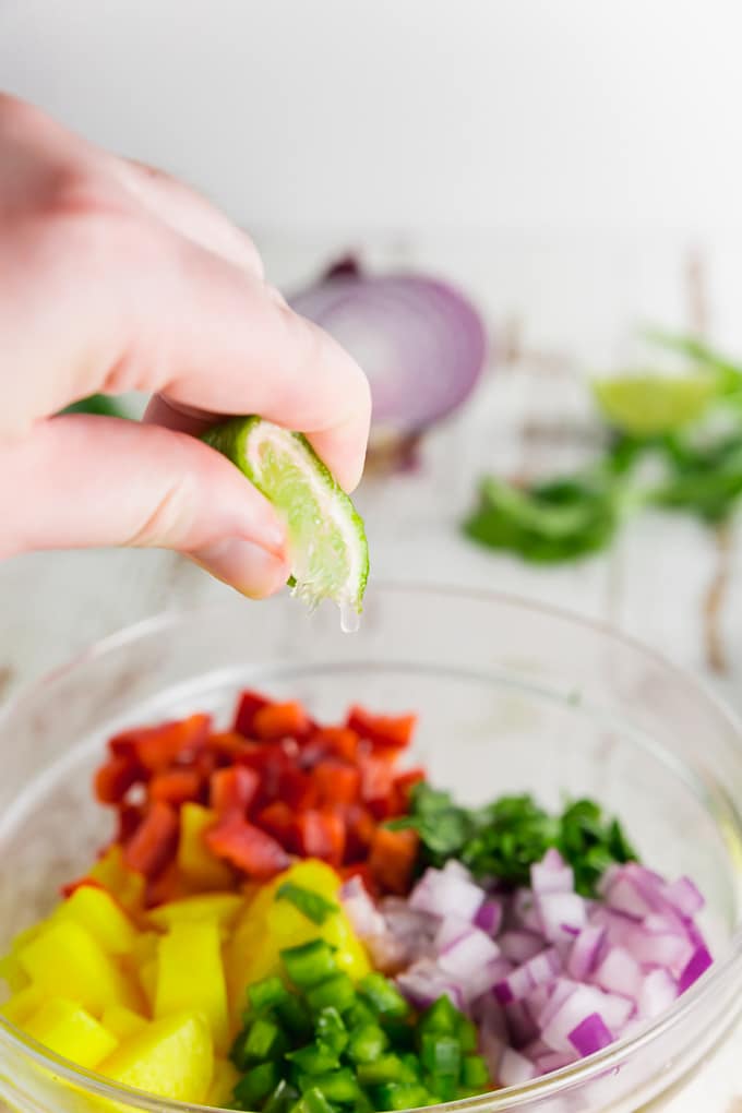 A photo of a hand squeezing a lime into a bowl of mango jalapeÃ±o salsa with red bell peppers.