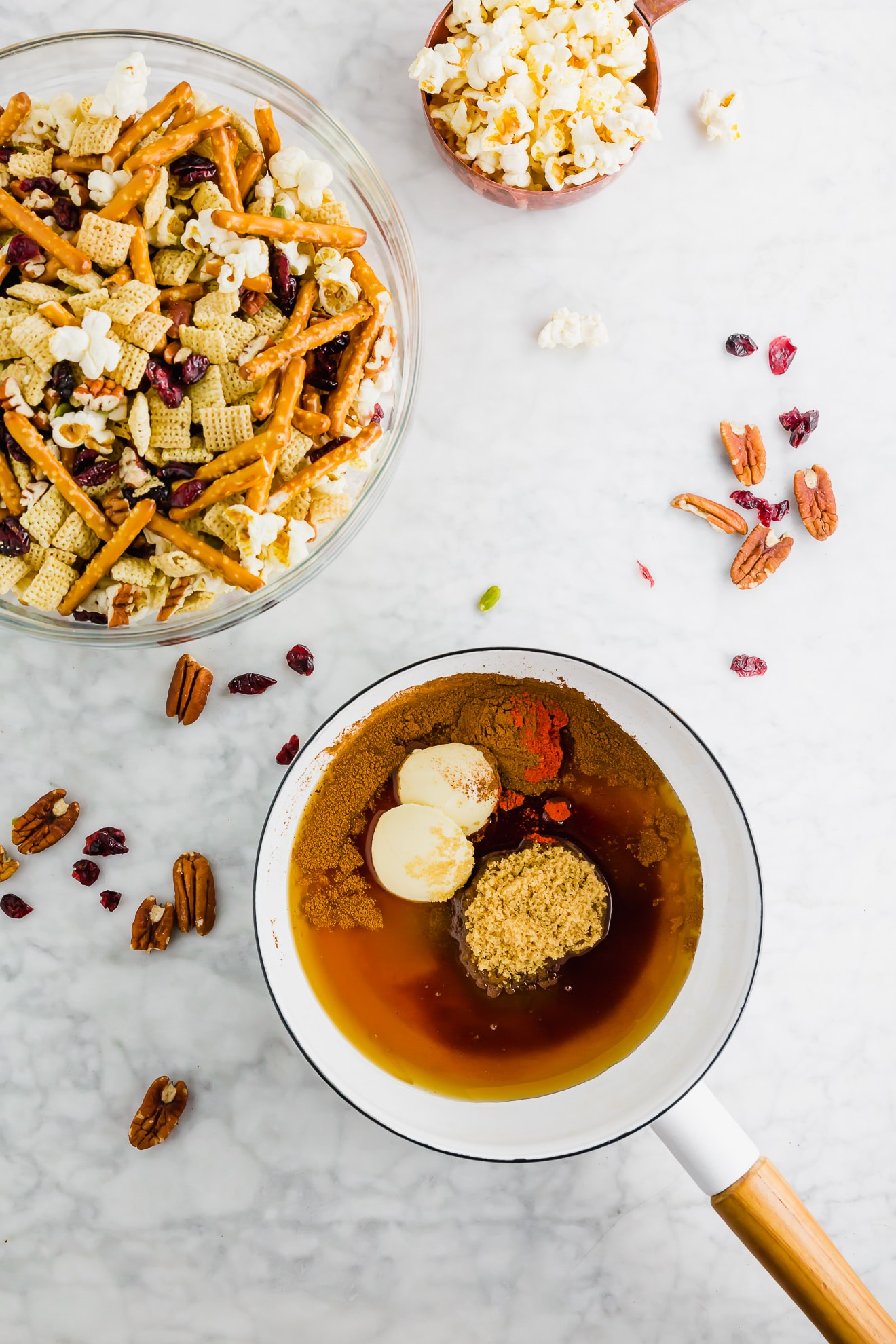 A photo of a saucepan with pure maple syrup, brown sugar, vegan butter, cinnamon, vanilla and cayenne pepper ready to be boiled with a bowl of gluten-free chex party mix in the corner.