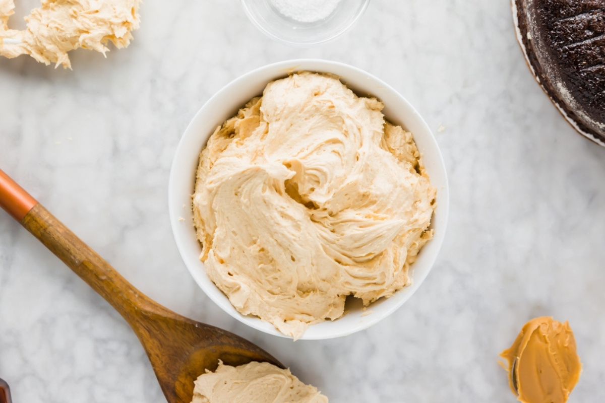 Overhead shot of a bowl of peanut butter frosting on marble with spoon and cake nearby.