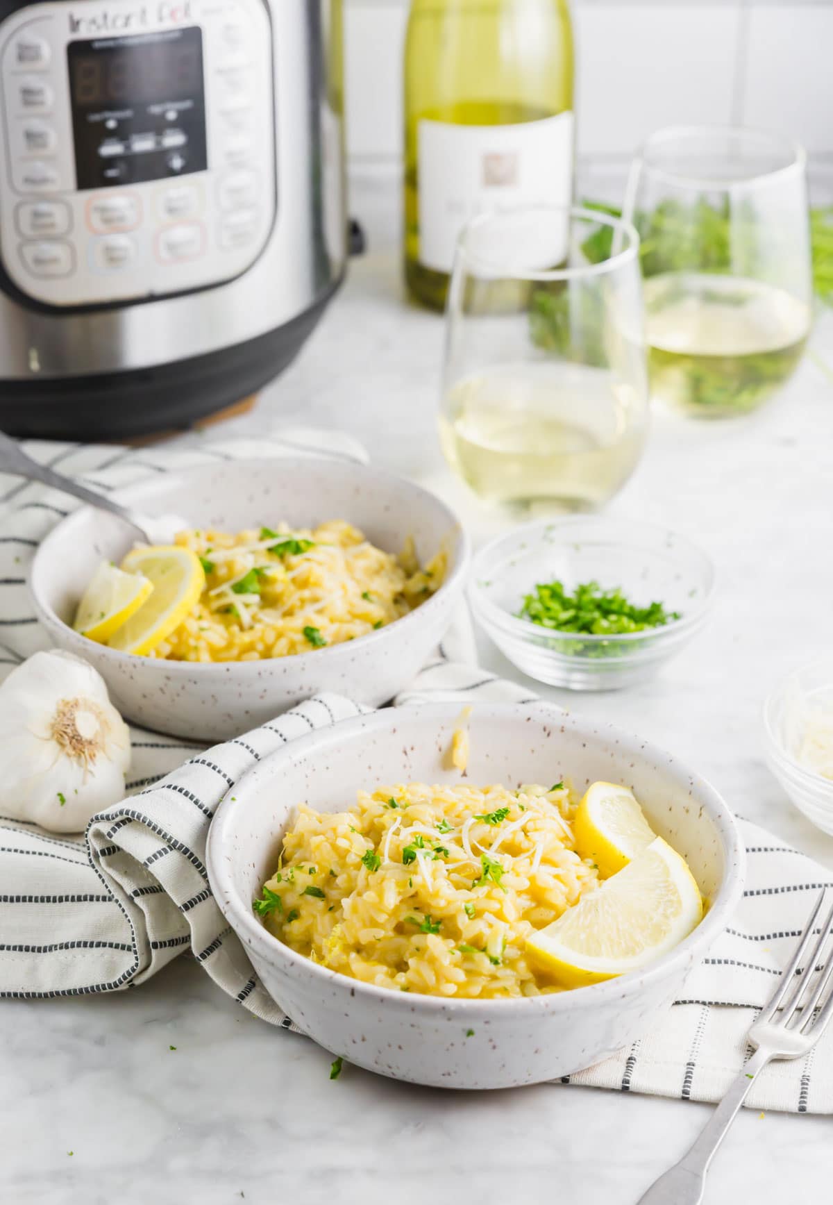A photo of two bowls of instant pot lemon risotto with an instant pot in the background with two glasses of wine.