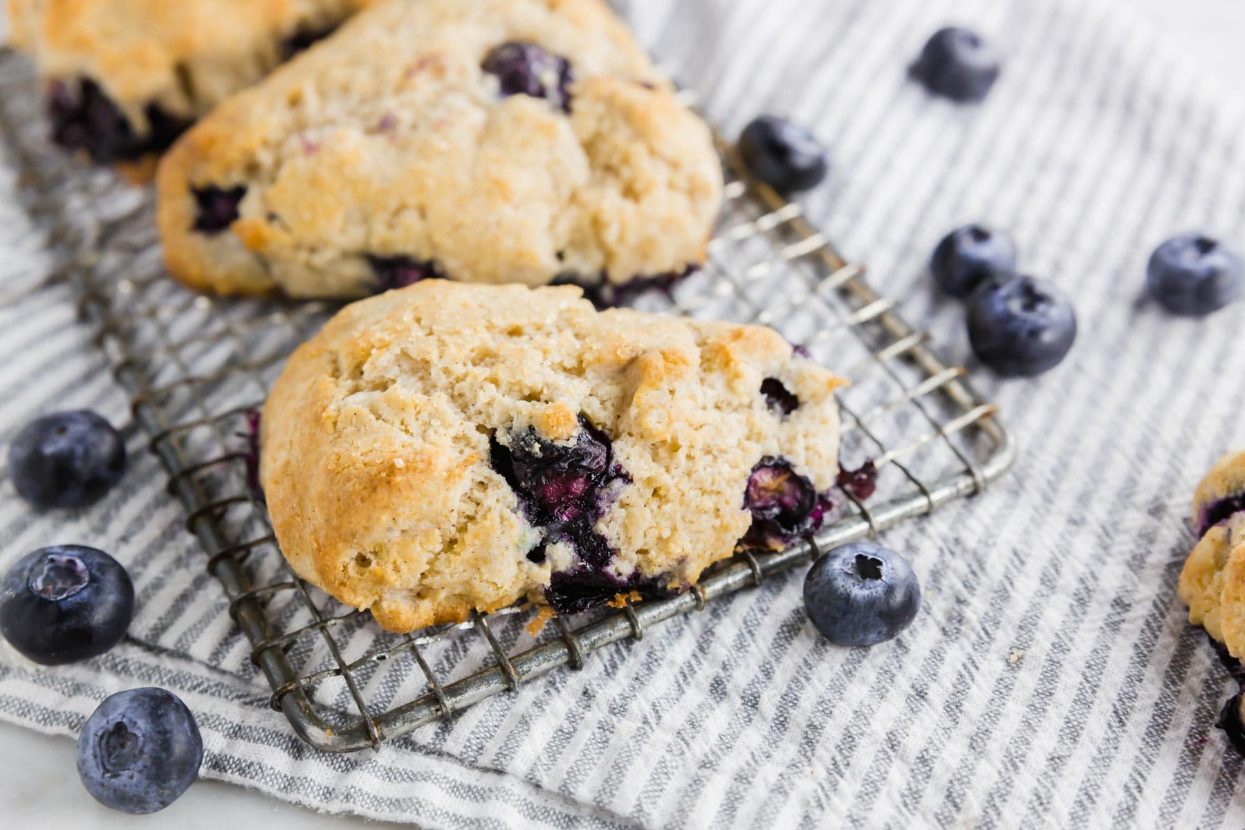 A close-up of a blueberry scone on a cooling rack, with fresh blueberries scattered on a striped cloth underneath.