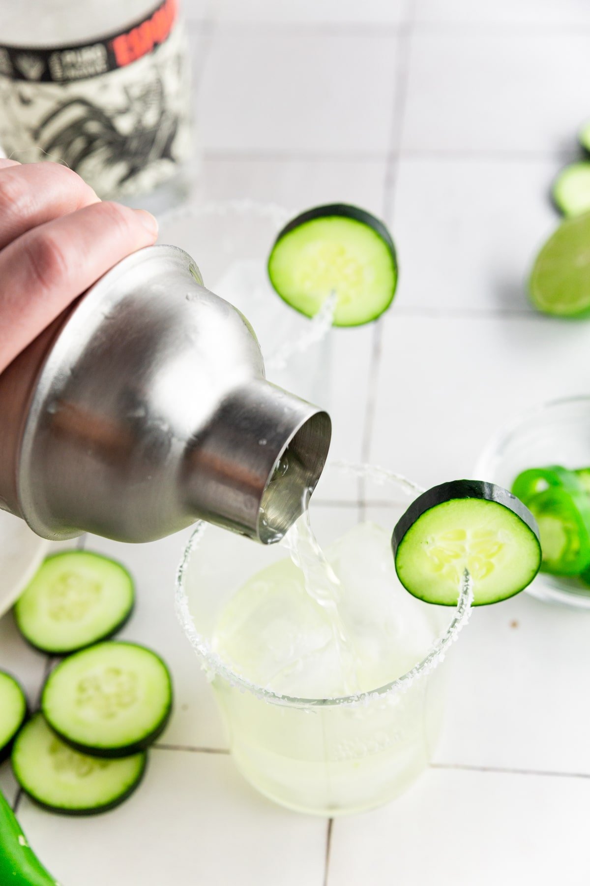 A photo of a cocktail shaker pouring cucumber jalapeño margarita into a glass rimmed with lime juice and kosher salt.