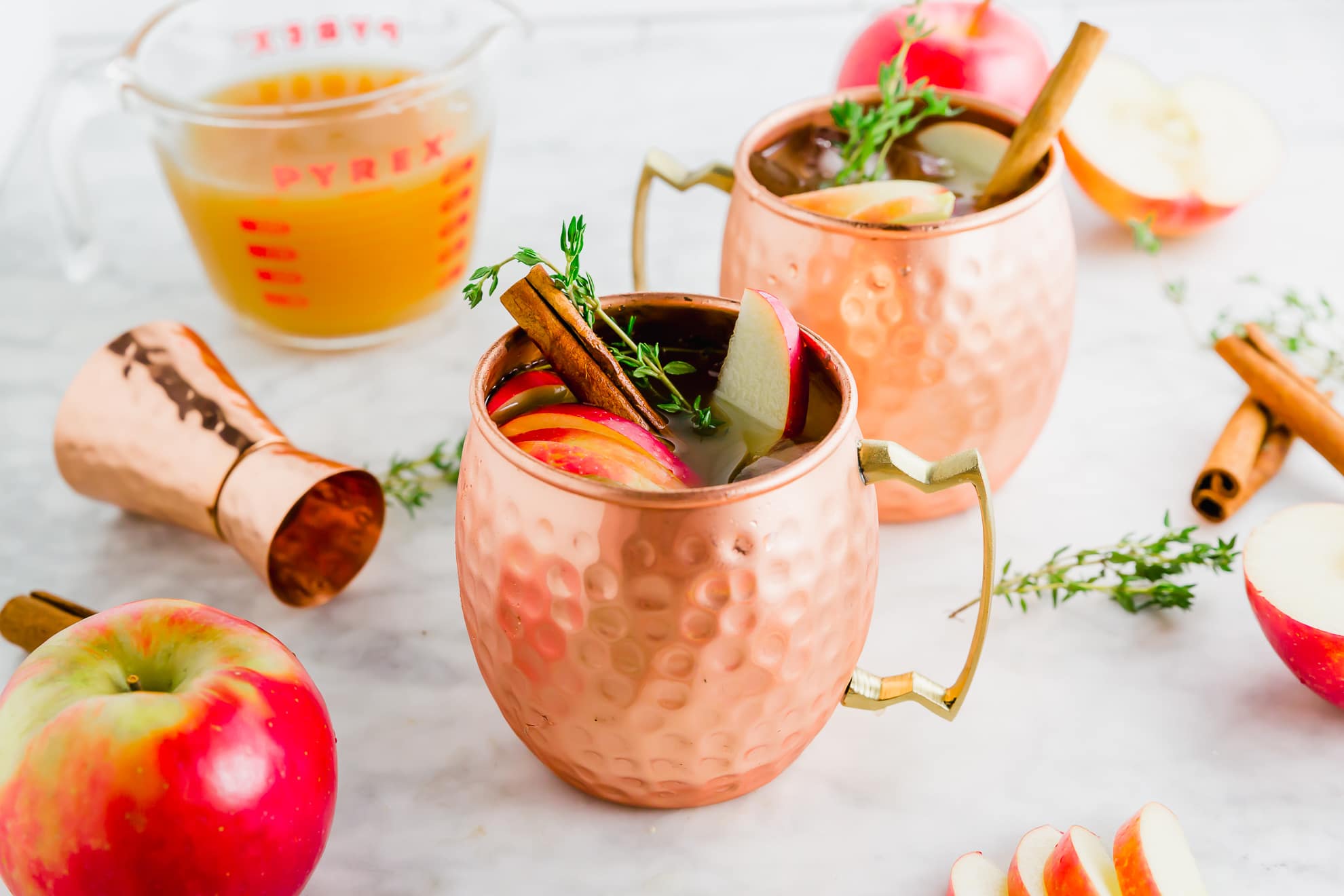 Two copper mugs filled with apple cider, garnished with apple slices, cinnamon sticks, and herbs, sit on a marble surface with fresh apples, cinnamon sticks, a jigger, and a measuring cup of cider in the background.
