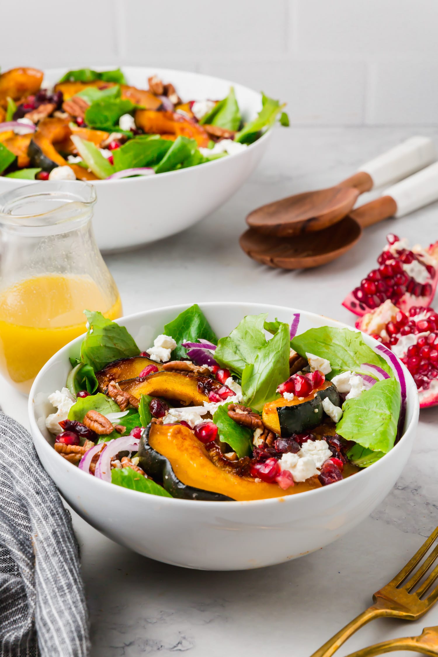 A photo of a bowl of fall harvest salad with acorn squash and pomegranate with a jar of apple cider vinaigrette and salad servers.