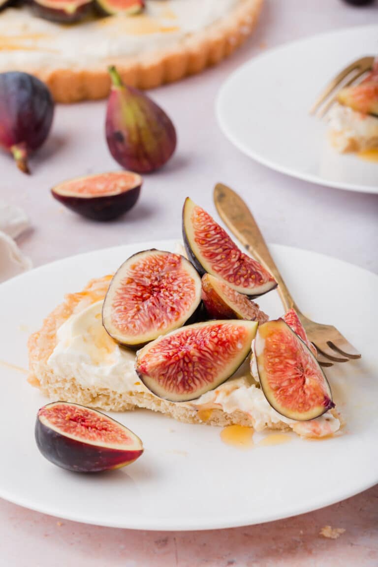 A slice of gluten-free fig tart with mascarpone cream and fresh fig wedges sits on a white plate with a fork. Whole and halved figs are scattered around, and another tart slice is visible in the background.