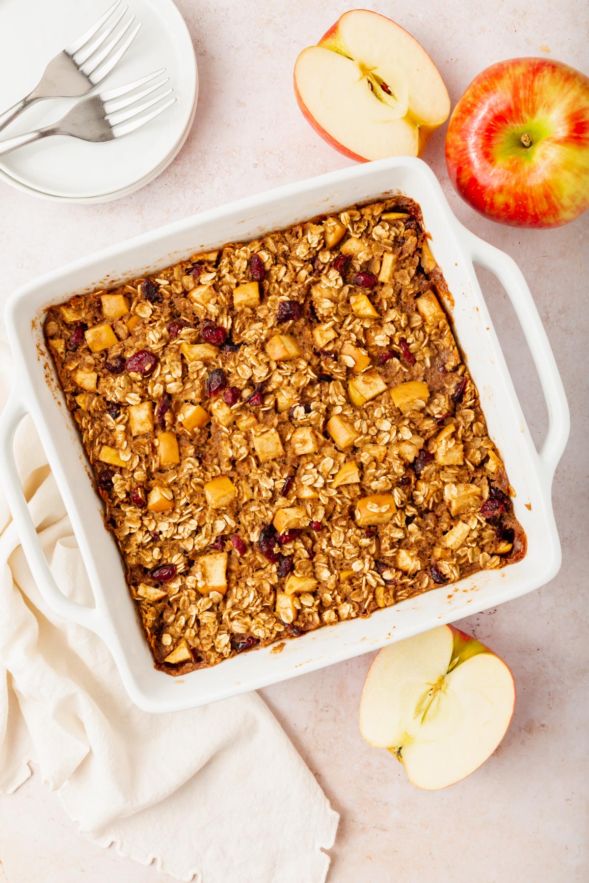 A white baking dish filled with apple cinnamon baked oatmeal containing apples and cranberries sits on a light surface. Sliced apples, a whole apple, a white napkin, and plates with forks are nearby.