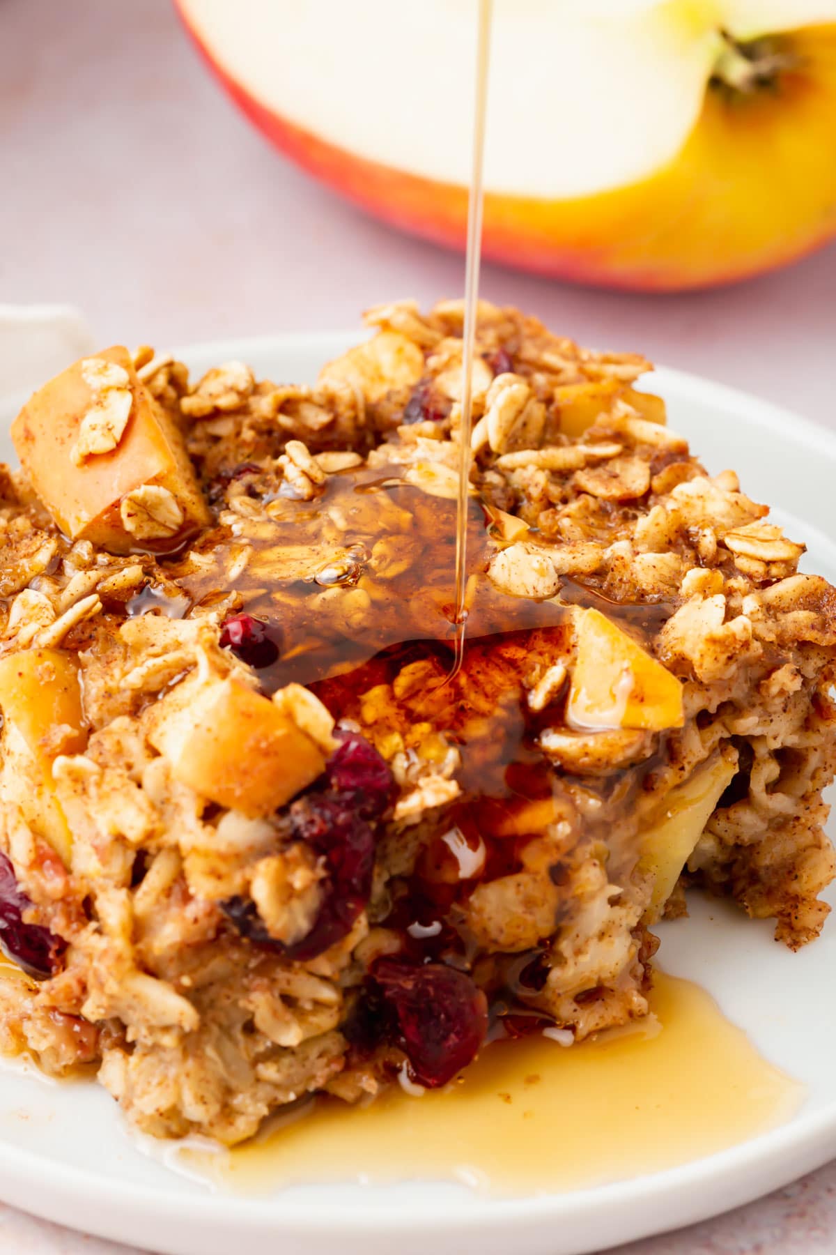 A slice of apple cinnamon baked oatmeal with apples and cranberries sits on a white plate, as syrup is being poured over it. An apple slice is blurred in the background.