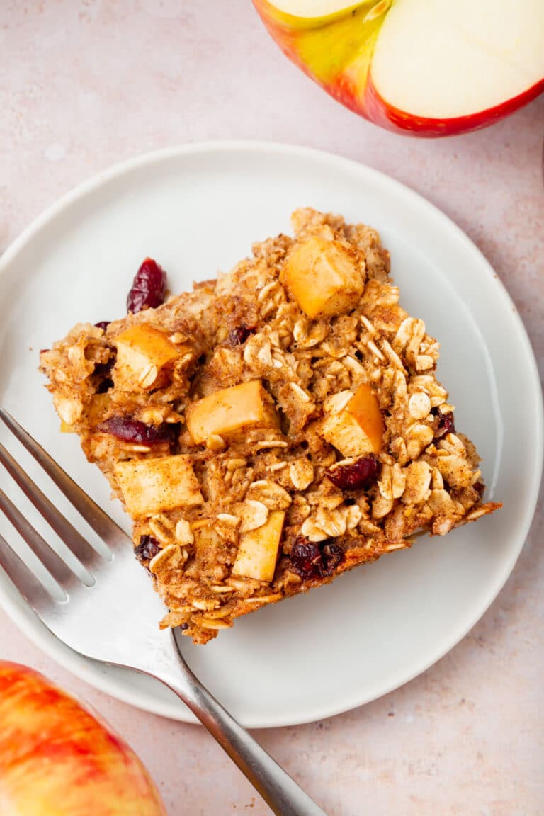 A square of apple cinnamon baked oatmeal with apple chunks and cranberries sits on a white plate with a fork. A sliced apple is visible nearby on a light pink surface.