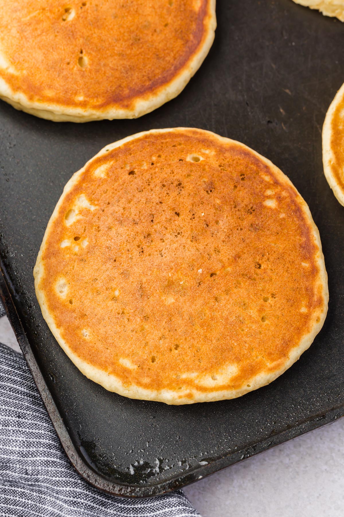 A close-up of a golden-brown gluten free pancake cooking on a griddle, with another pancake partially visible nearby. A gray and white striped cloth is at the edge of the griddle.