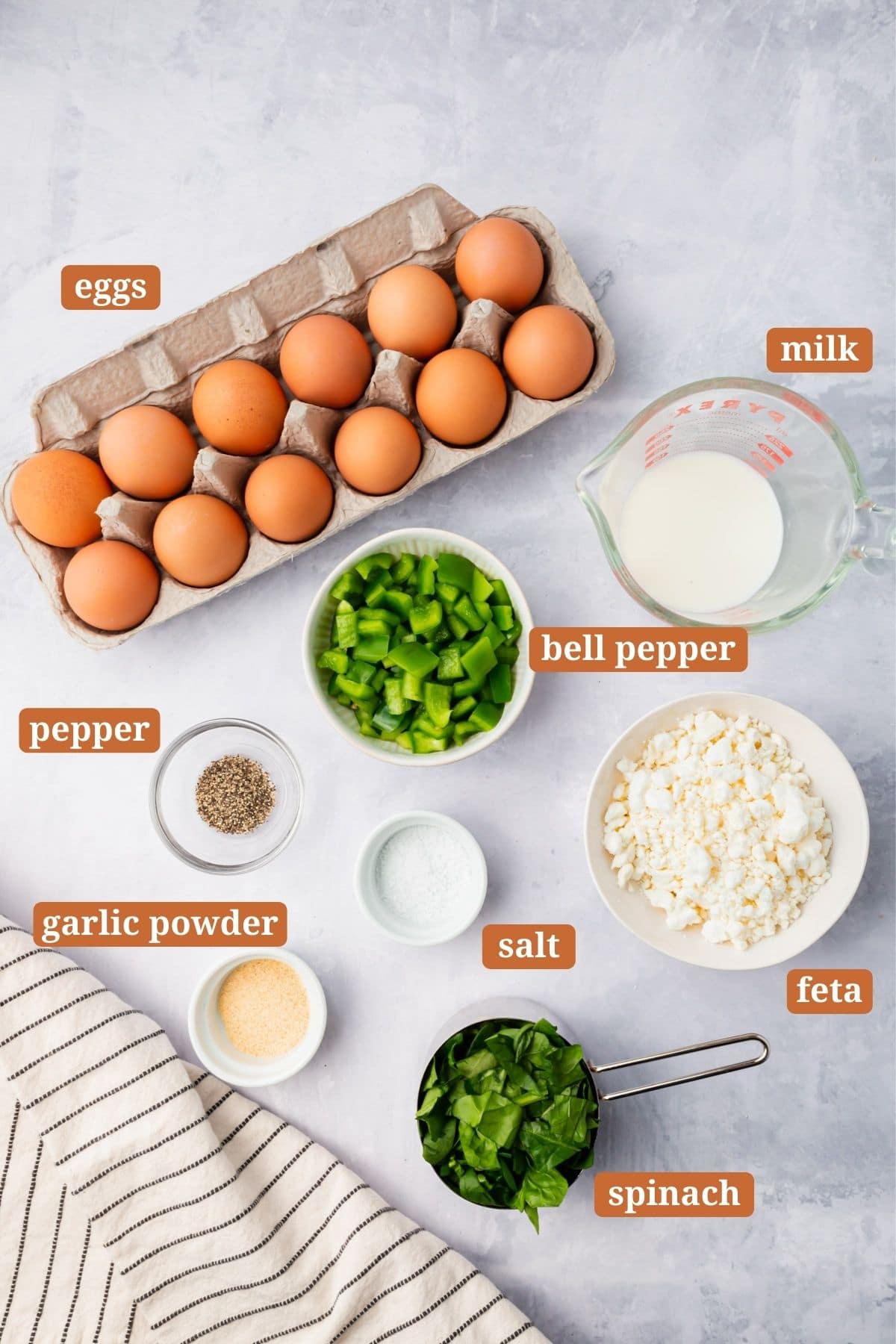 Top-down view of labeled ingredients for spinach feta egg muffins on a light surface: a carton of eggs, milk, green bell pepper, pepper, garlic powder, salt, feta cheese, and chopped spinach beside a striped towel.