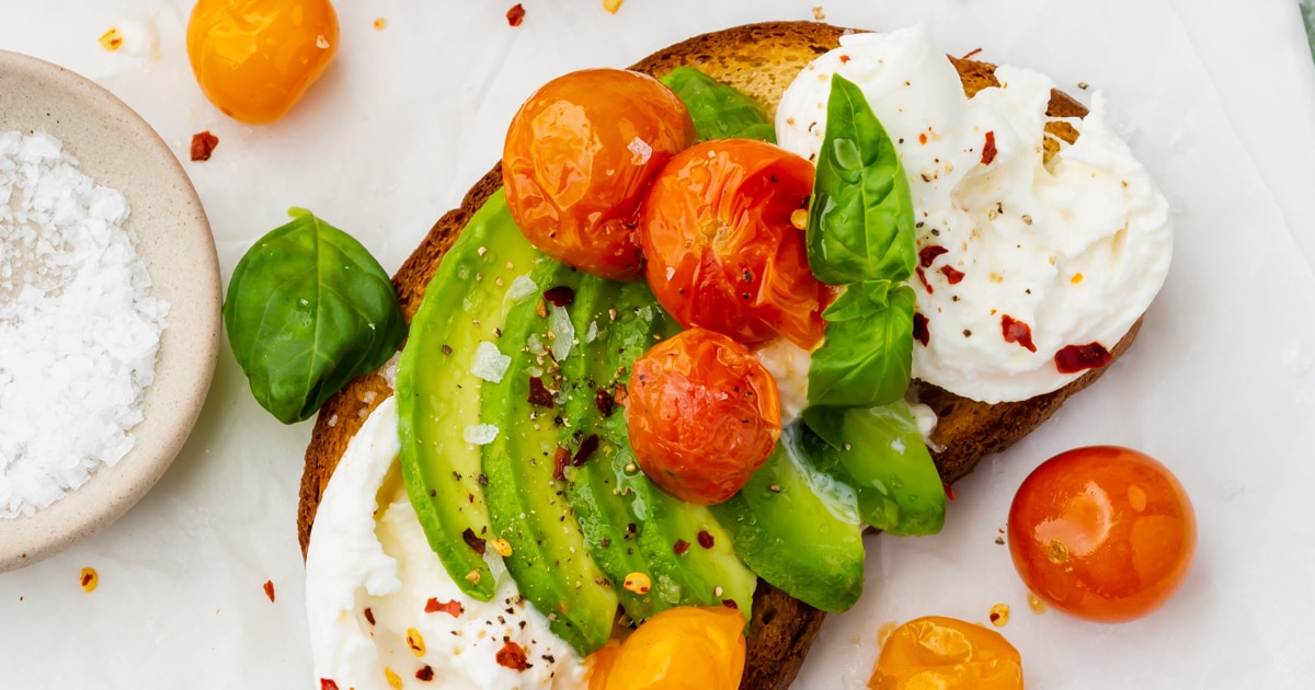 A closeup of avocado burrata toast topped with roasted tomatoes next to a bowl of flaky sea salt.