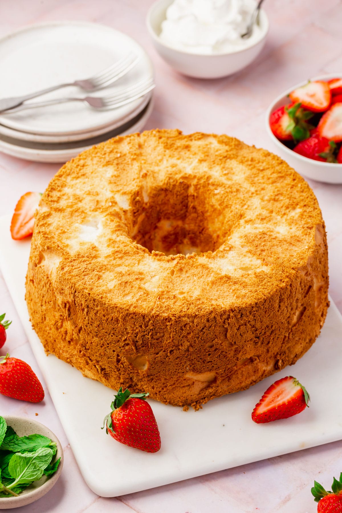An angel food cake on a serving platter with strawberries scattered around, plates and forks in the background and small bowls of whipped topping, strawberries and mint leaves.