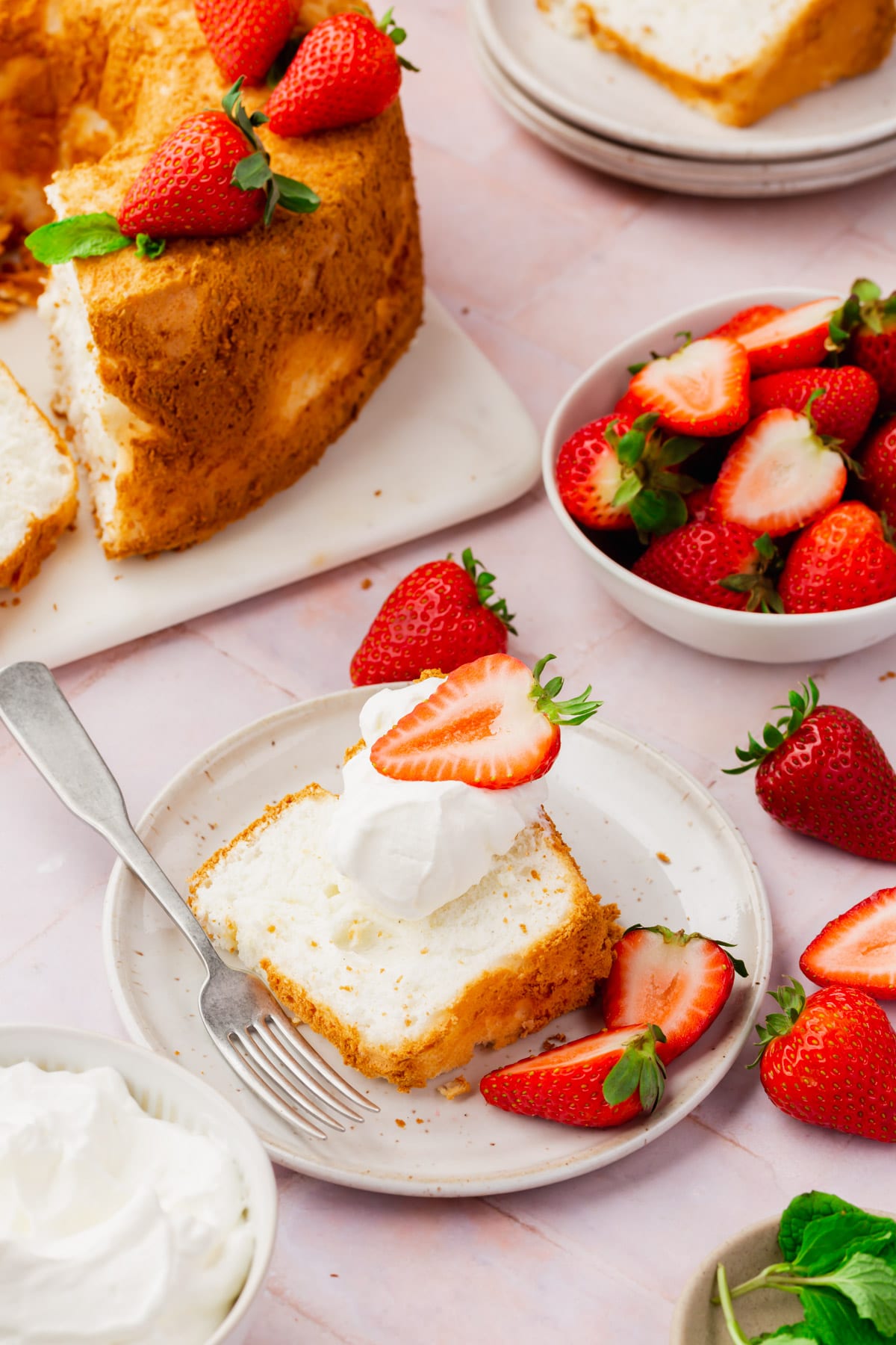 A slice of angel food cake on a plate topped with whipped cream and a strawberry with a full angel food cake and a bowl of strawberries in the background.