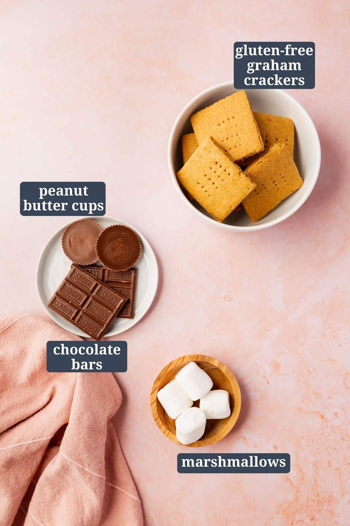 An overhead shot of three bowls filled with gluten-free graham crackers, peanut butter cups and hershey bars in another bowl, and marshmallows in the last bowl.