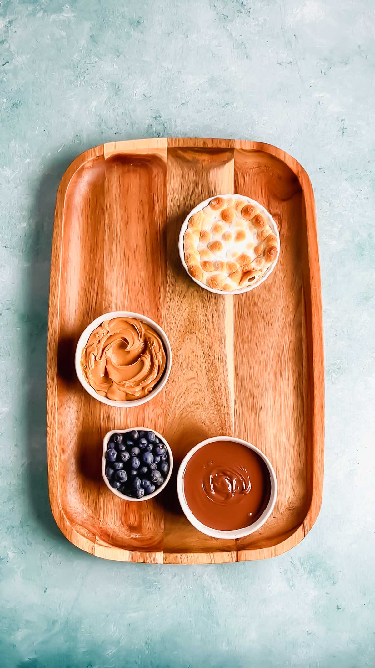 A wood rectangular charcuterie board with small bowls of s'mores dip, peanut butter, nutella, and blueberries.