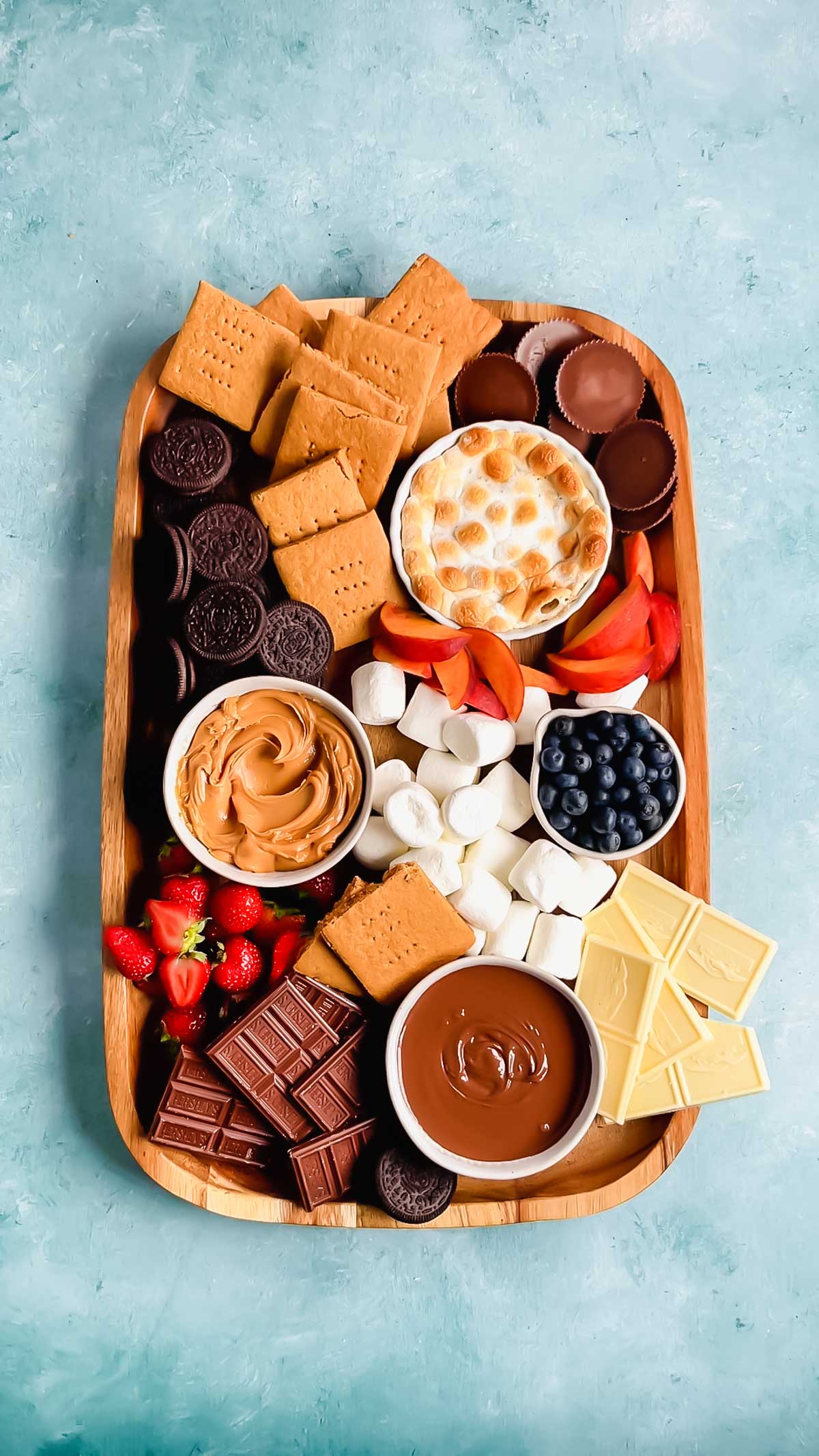 A wooden board with graham crackers, oreos, peanut butter cups, peaches, marshmallows, chocolate bars, strawberries, and bowls of peanut butter, blueberries, s'mores dip, and nutella.