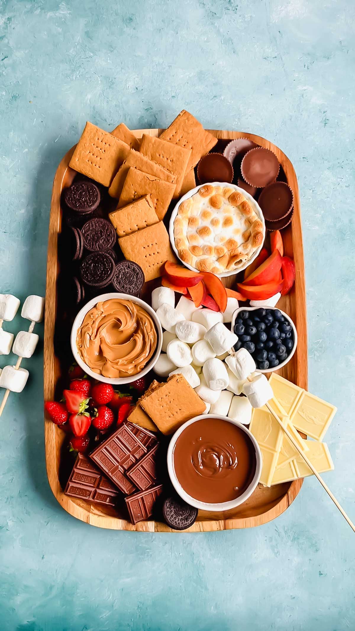 A wood board with graham crackers, oreos, marshmallows, chocolate bars, strawberries, peanut butter cups, peaches, and bowls of blueberries, nutella, peanut butter, and s'mores dip with a few marshmallows on skewers.