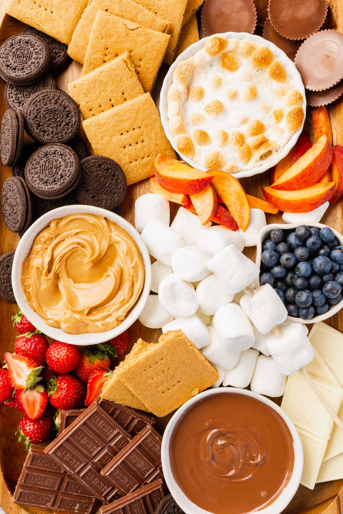 A s'mores board filled with gluten-free oreos, graham crackers, s'mores dip, peaches, blueberries, marshmallows, peanut butter, nutella, strawberries, white chocolate, and marshmallows.