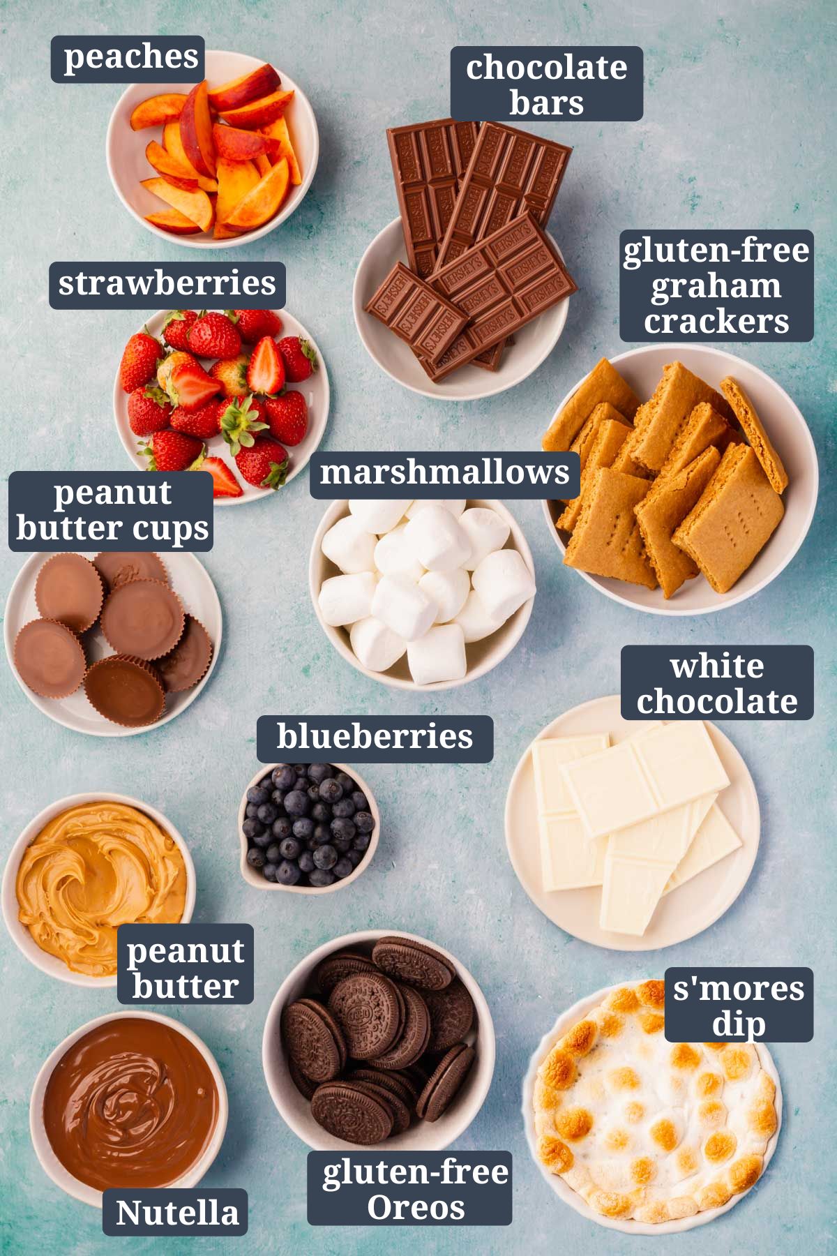 Small bowls of ingredients to make s'mores board on a blue table.