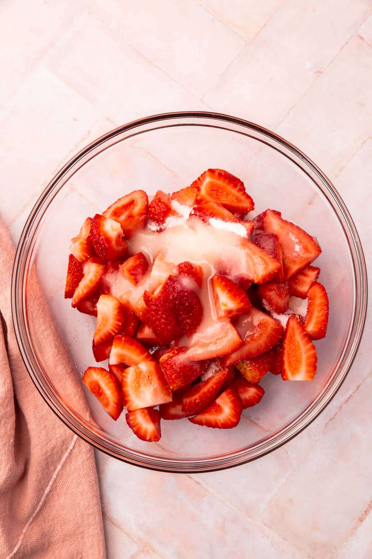 A glass mixing bowl with sliced strawberries, granulated sugar, and lemon juice in it.