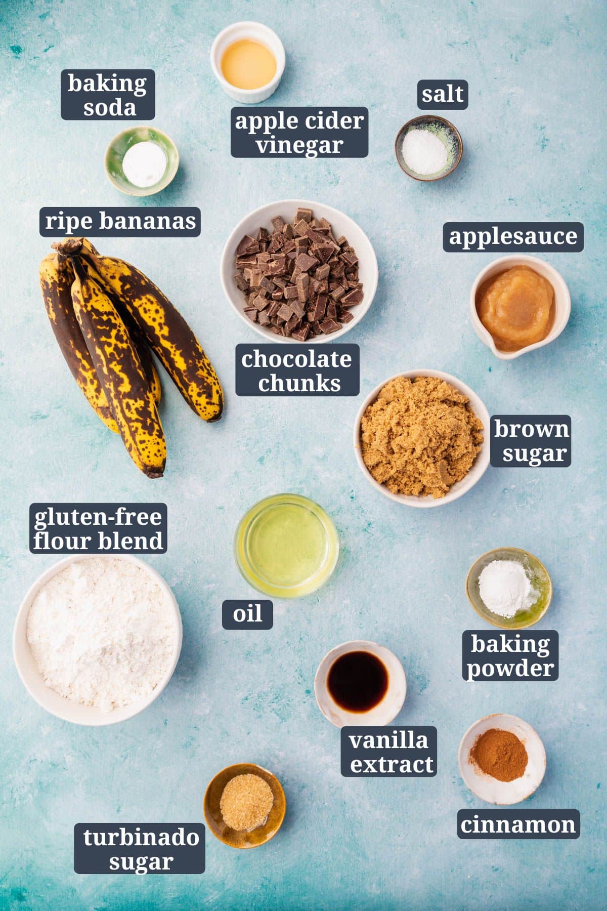 Ingredients in small bowls on a blue background to make gluten-free vegan chocolate chunk banana bread, including very ripe bananas, chocolate chunks, brown sugar, applesauce, vanilla, cinnamon, oil, turbinado sugar, and gluten-free flour blend.