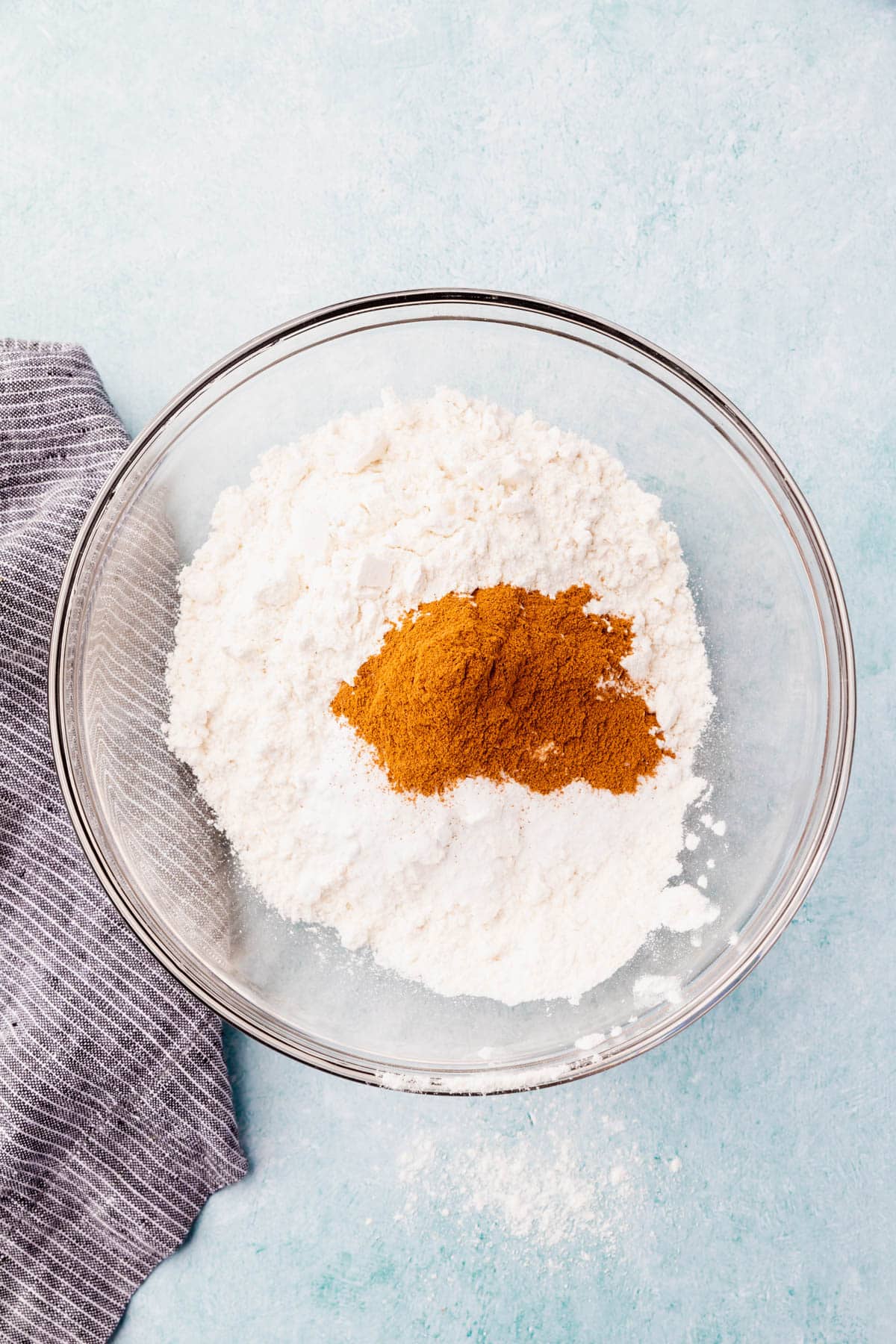 A glass mixing bowl on a blue table with gluten-free flour, cinnamon, baking powder, baking soda, and salt before mixing together.