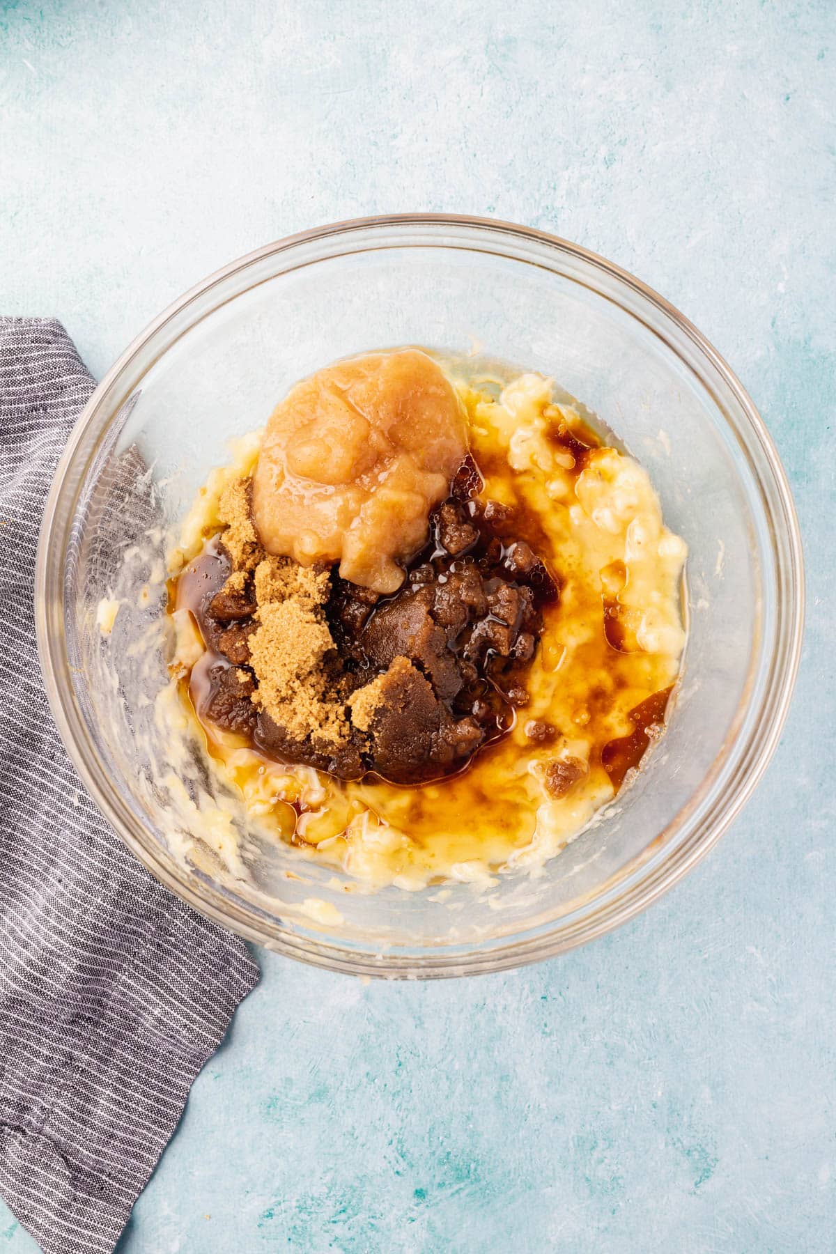 A glass mixing bowl on a blue table with mashed banana, brown sugar, oil, applesauce, vanilla, and apple cider vinegar before mixing.