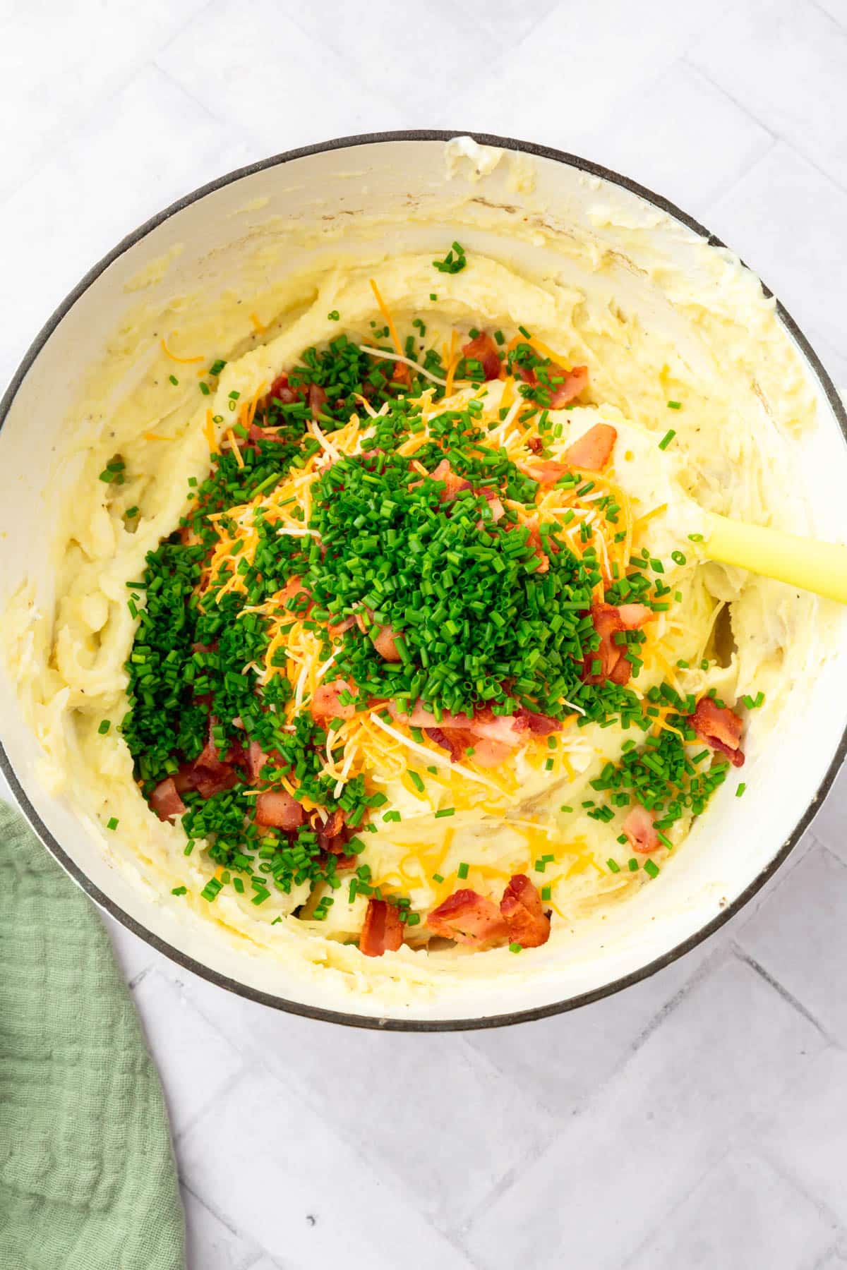 A large dutch oven filled with mashed potatoes topped with chopped chives, cooked bacon bits and cheddar cheese before mixing together.