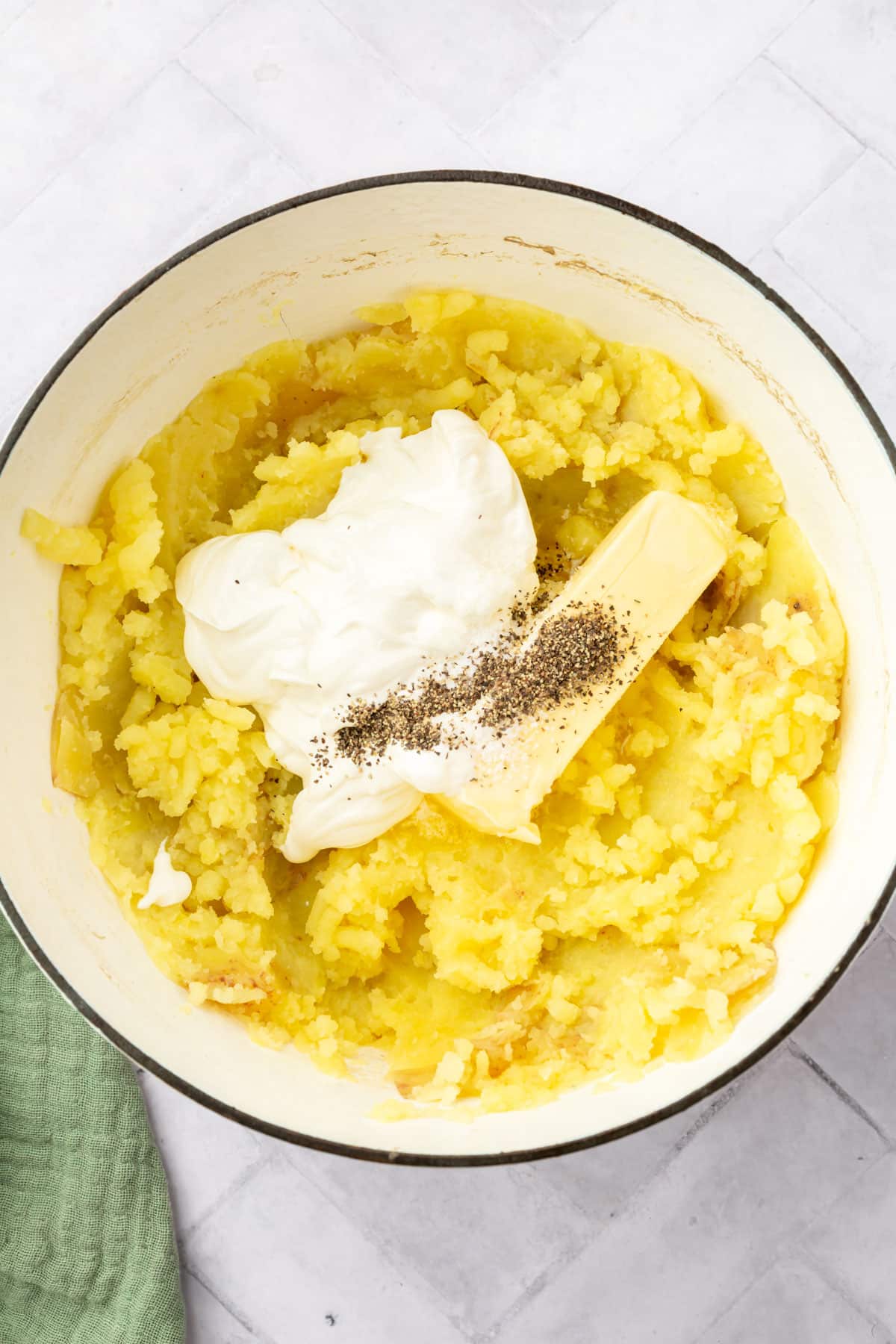 A large dutch oven filled with mashed potatoes topped with sour cream, salt, black pepper and a stick of butter before mixing together.