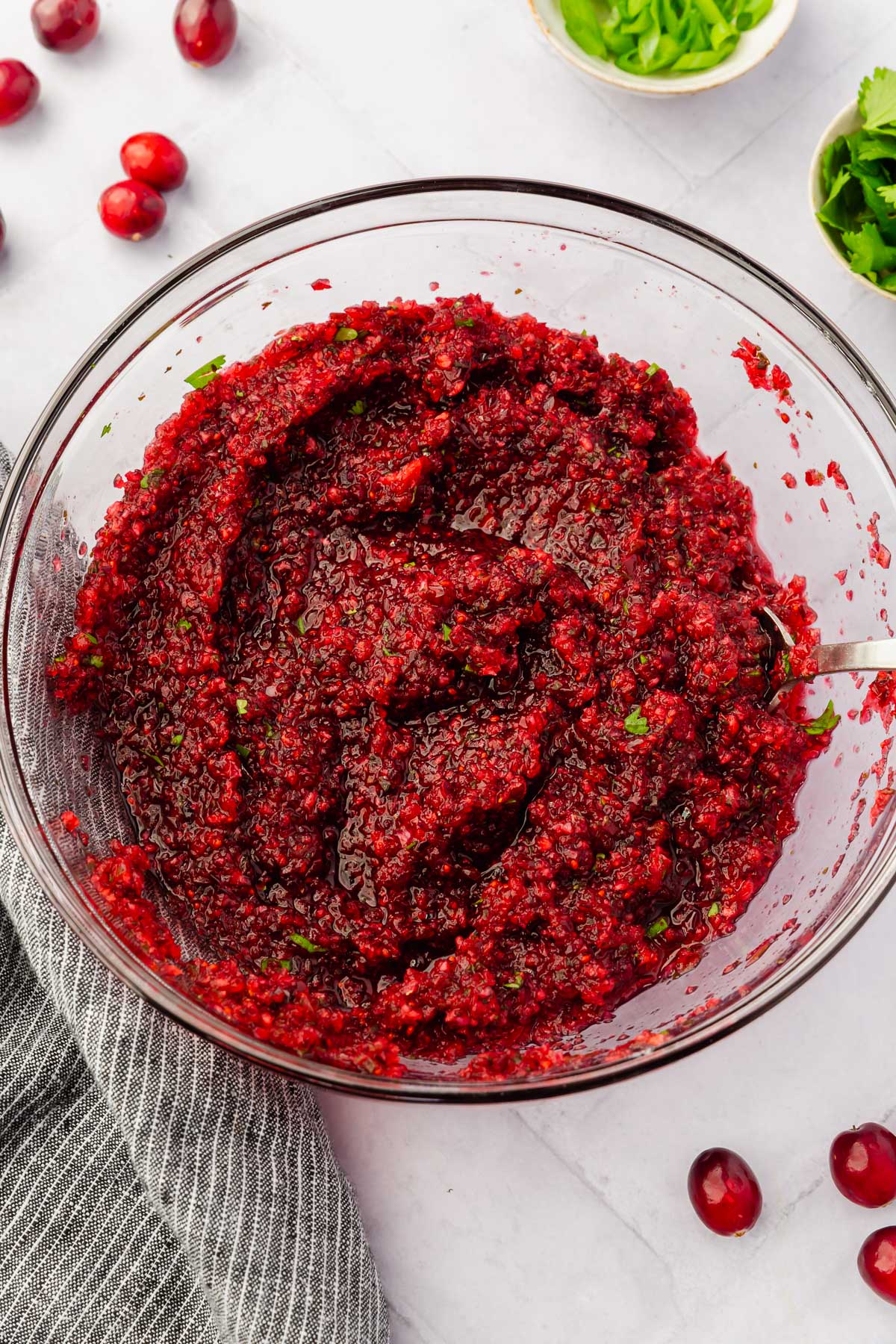 A clear mixing bowl with cranberry salsa in it with bowls of green onions and cilantro on the side.