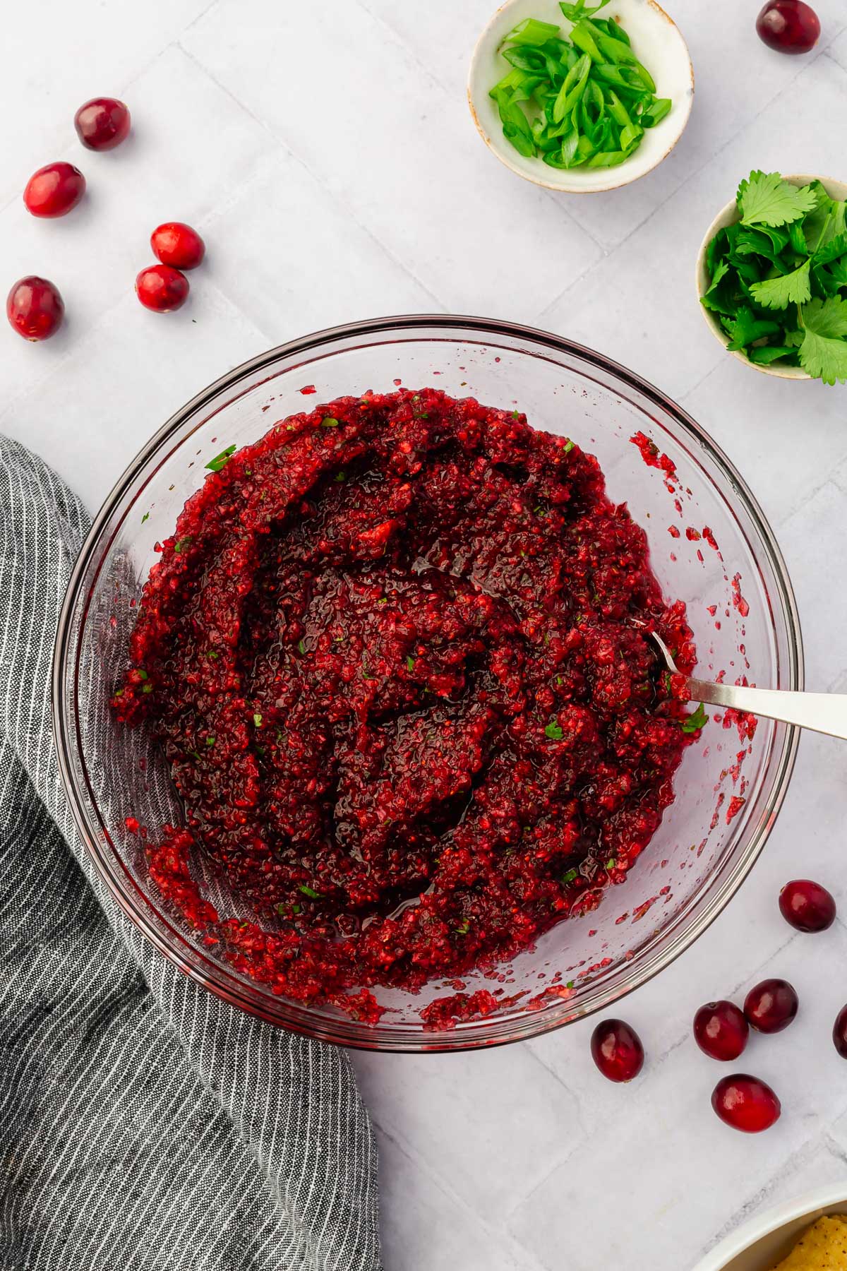 A clear mixing bowl with cranberry salsa in it with bowls of green onions and cilantro on the side.