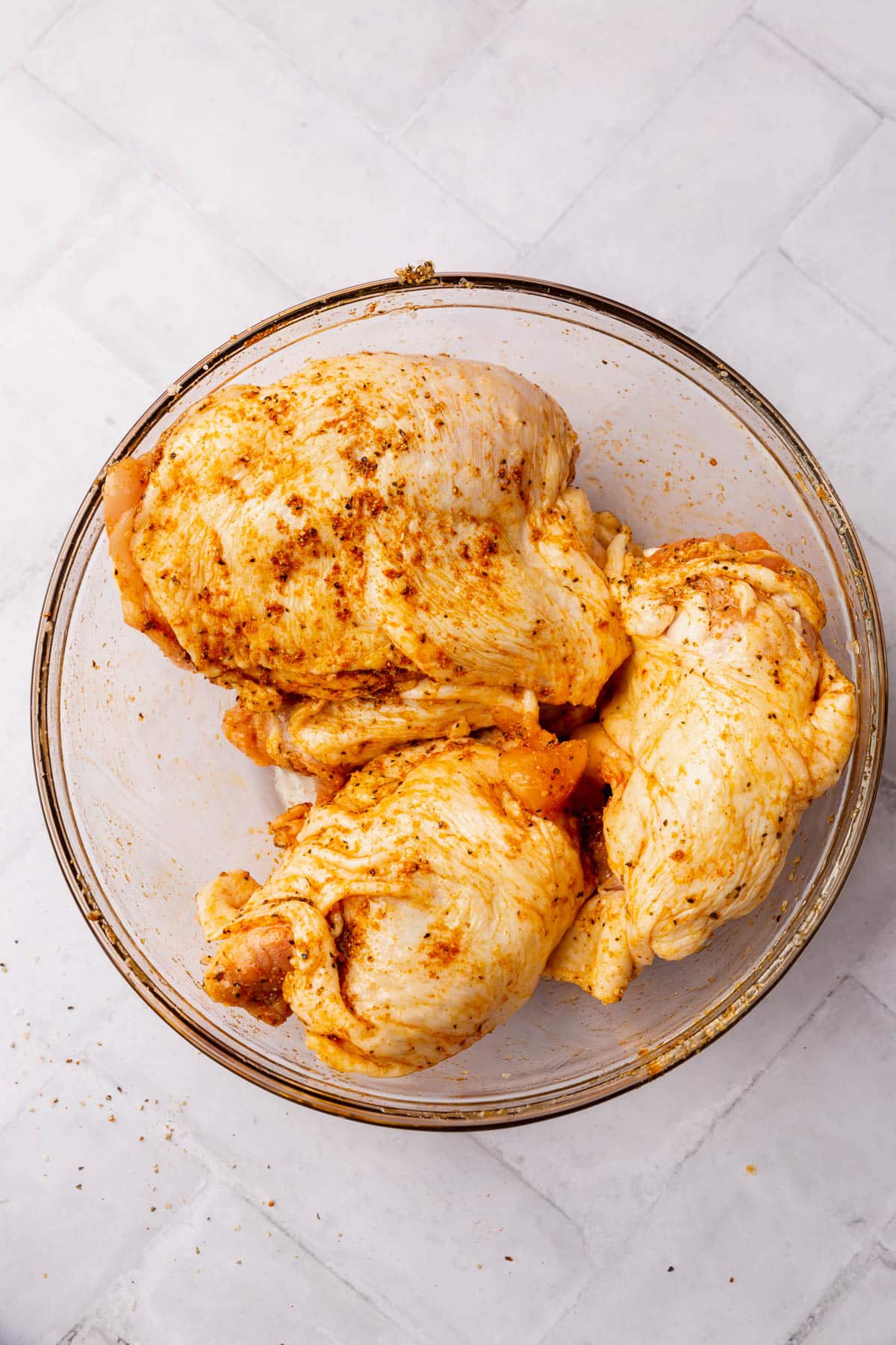 Raw chicken thighs that have been covered in oil and a seasoning blend in a glass mixing bowl.