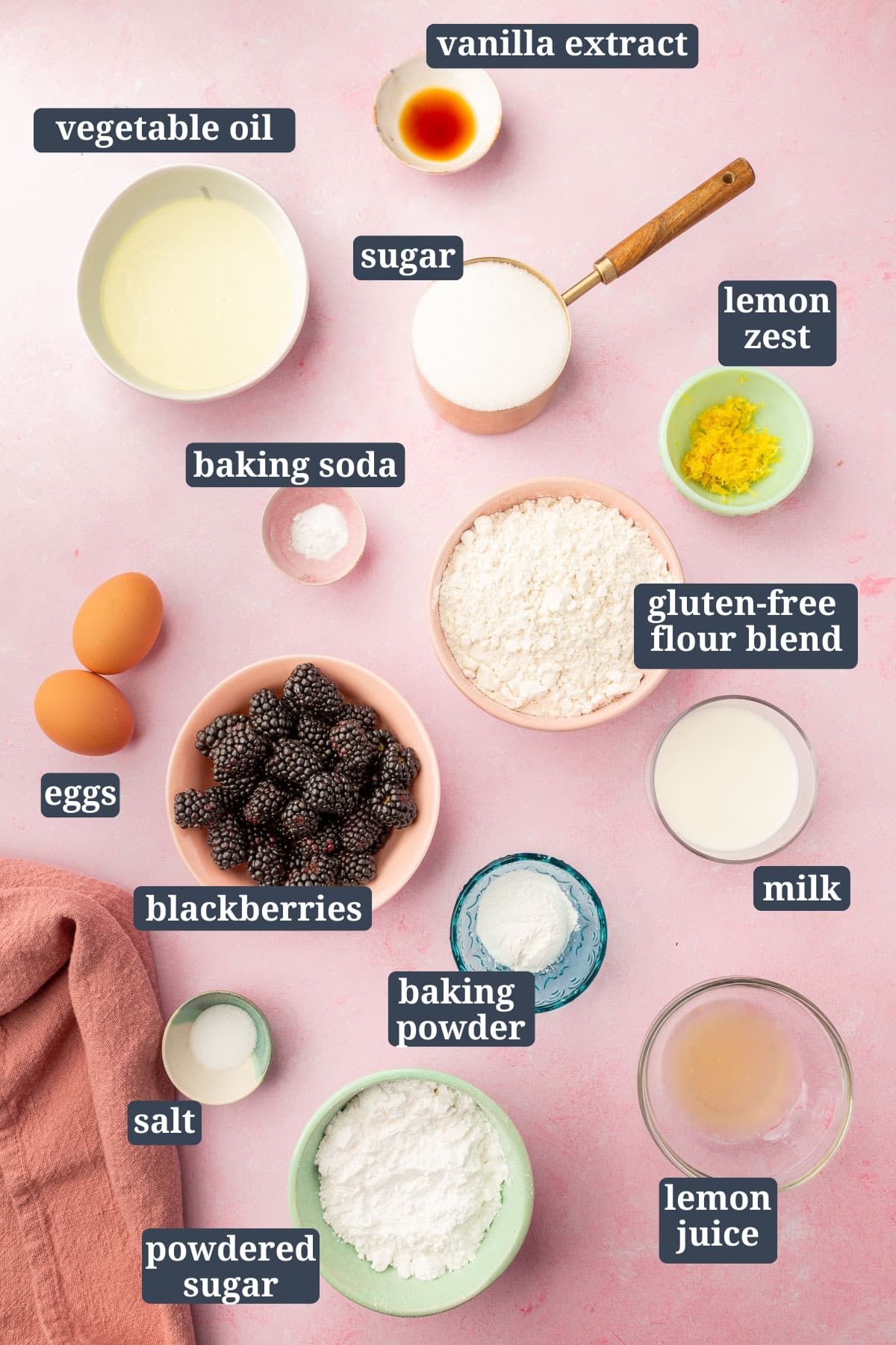 An overhead view of small bowls of ingredients to make gluten free blackberry muffins, including vegetable oil, vanilla, sugar, lemon zest, baking soda, gluten-free flour blend, eggs, baking powder, milk, lemon juice, powdered sugar, salt, and blackberries with text overlays over each ingredient.