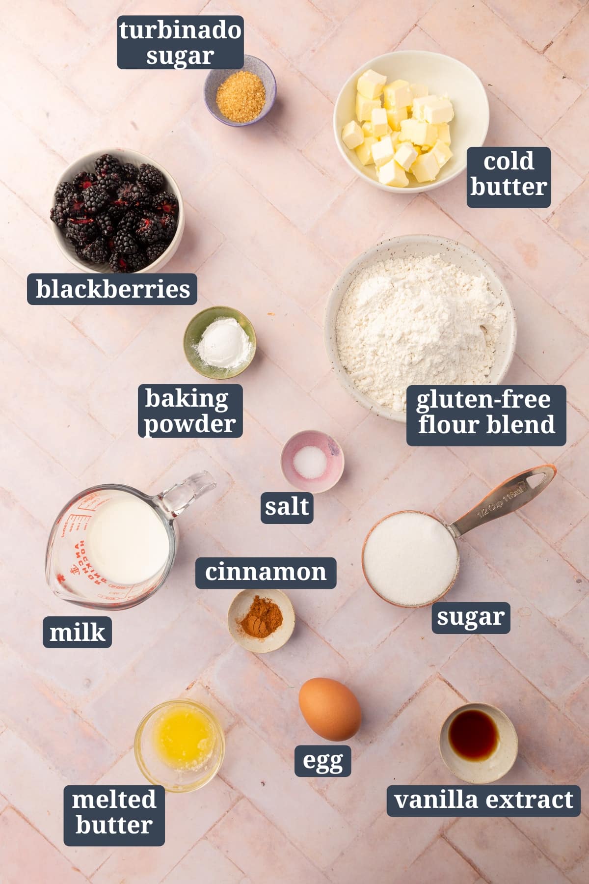Flat lay of ingredients for Gluten-Free Blackberry Scones, each labeled: turbinado sugar, cold butter, blackberries, baking powder, gluten-free flour blend, sugar, salt, cinnamon, milk, melted butter, an egg, and vanilla extract on a light pink surface.