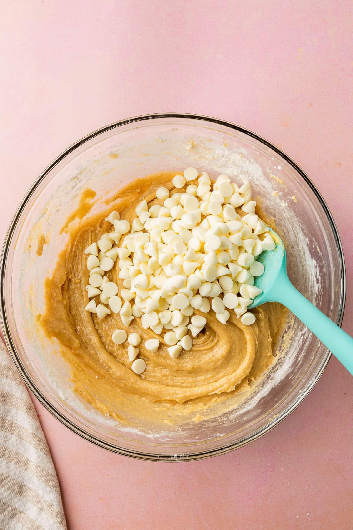 A glass mixing bowl with gluten-free blondie batter topped with a heaping pile of white chocolate chips and a blue spatula.