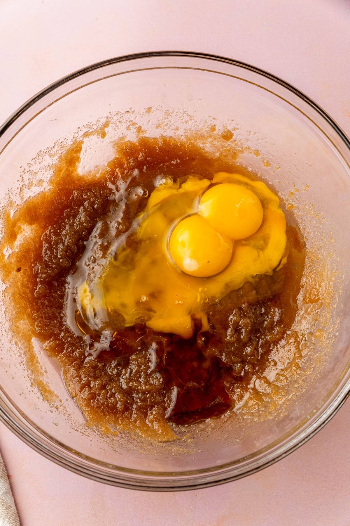 A glass mixing bowl filled with a brown sugar and melted butter mixture topped with eggs and vanilla extract.