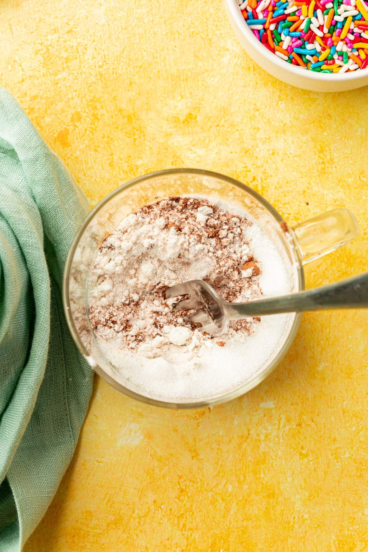 Gluten-free flour, cocoa powder, salt, and granulated sugar being mixed together by a fork in a glass mug.