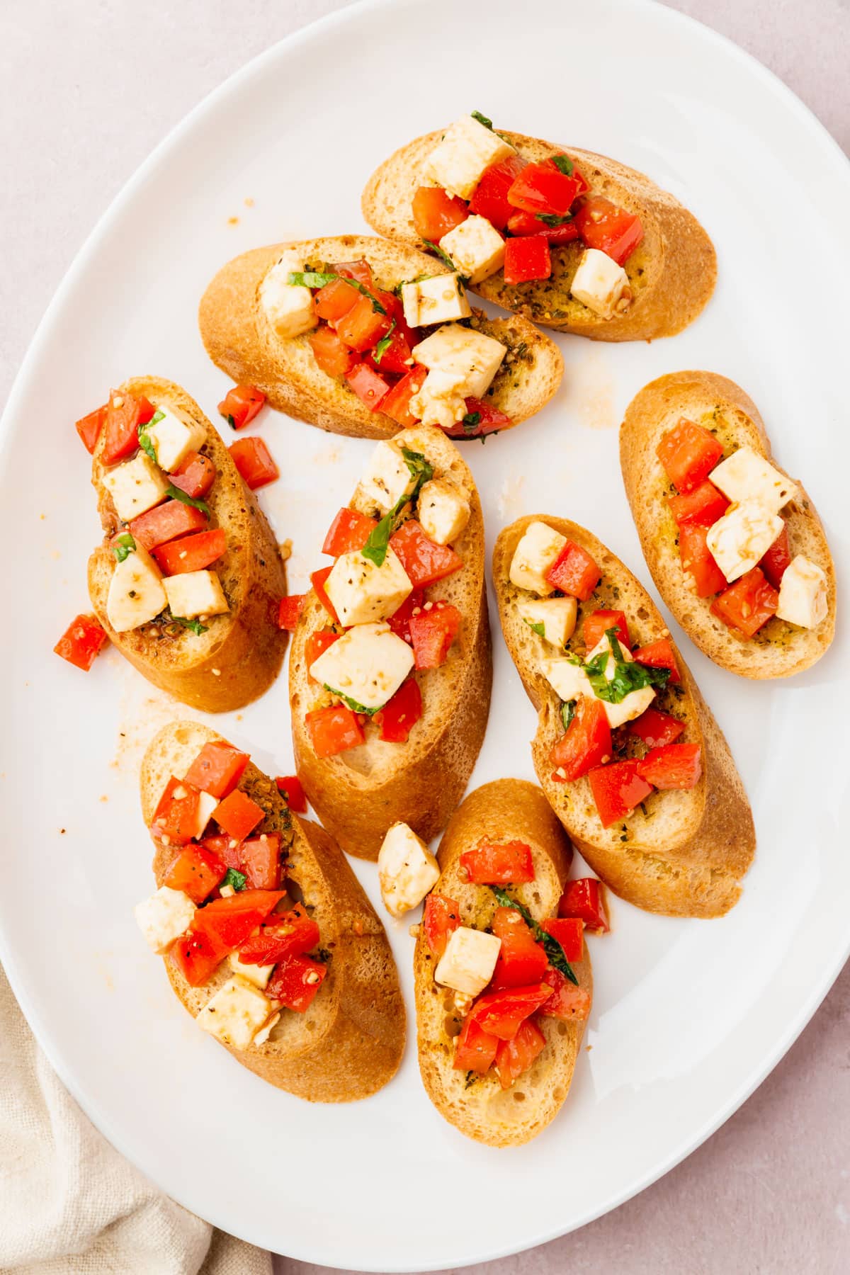 Eight pieces of gluten-free bread topped with diced tomatoes, diced mozzarella and basil on a white oval platter.