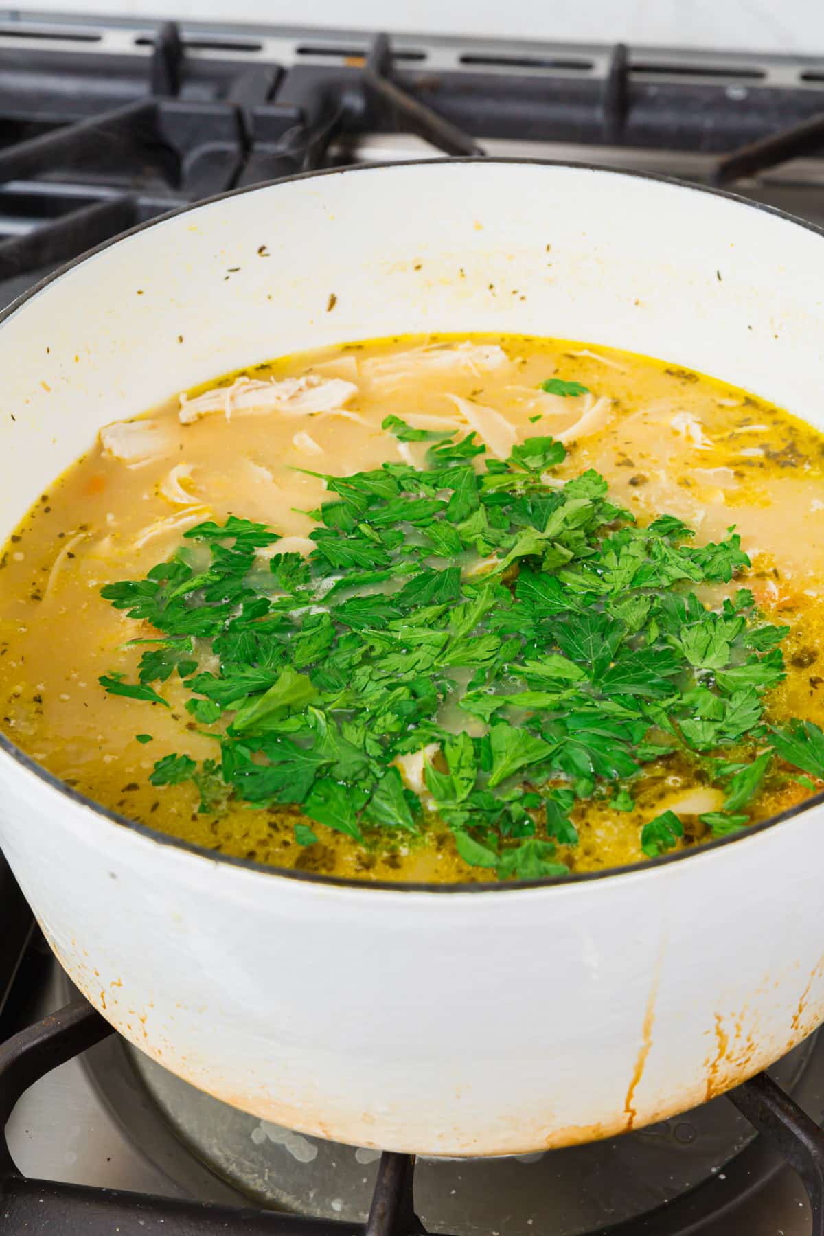 A white dutch oven on the stove with gluten free chicken noodle soup in it topped with a pile of chopped fresh parsley before mixing together.