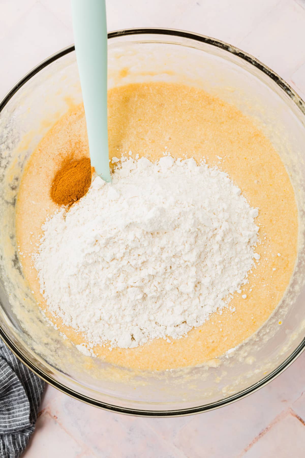 A glass mixing bowl with banana bread wet ingredients topped with gluten free flour and ground cinnamon before mixing together.