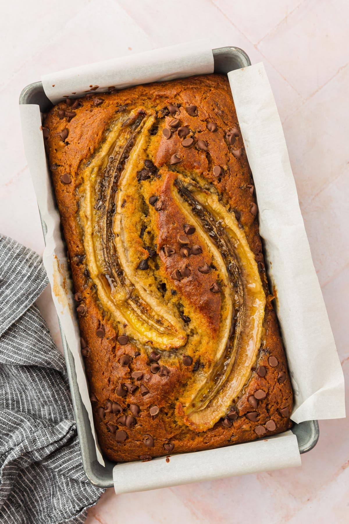 Gluten-free banana bread with chocolate chips and large banana slices on top in a loaf pan lined with parchment paper.