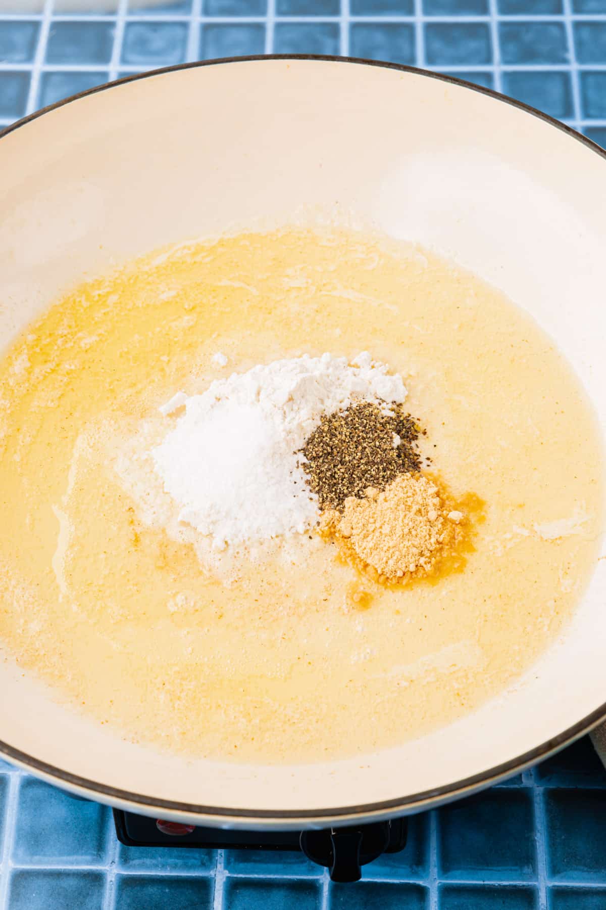 A white braising pan with melted butter, gluten-free flour, ground mustard powder and black pepper before mixing together to make a roux.