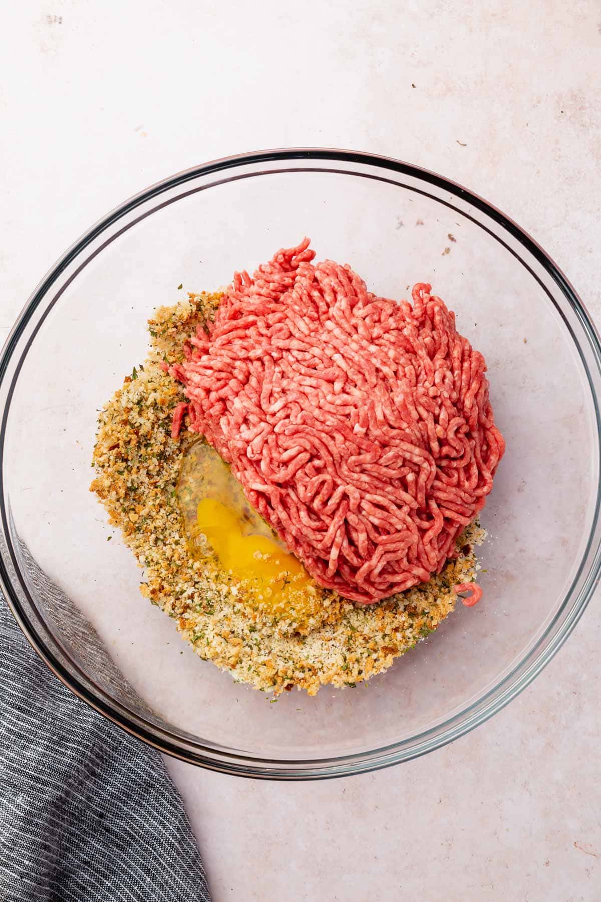 A glass mixing bowl with ground beef, egg, and a gluten-free breadcrumb mixture to make meatballs that are gluten-free.