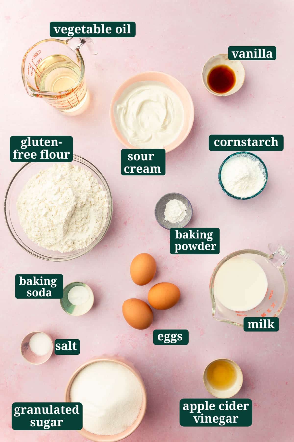 Ingredients in small bowls to make gluten-free vanilla cake including oil, sour cream, vanilla, gluten-free flour, baking powder, cornstarch, eggs, salt, baking soda, granulated sugar, apple cider vinegar and milk with text overlays over each ingredient.