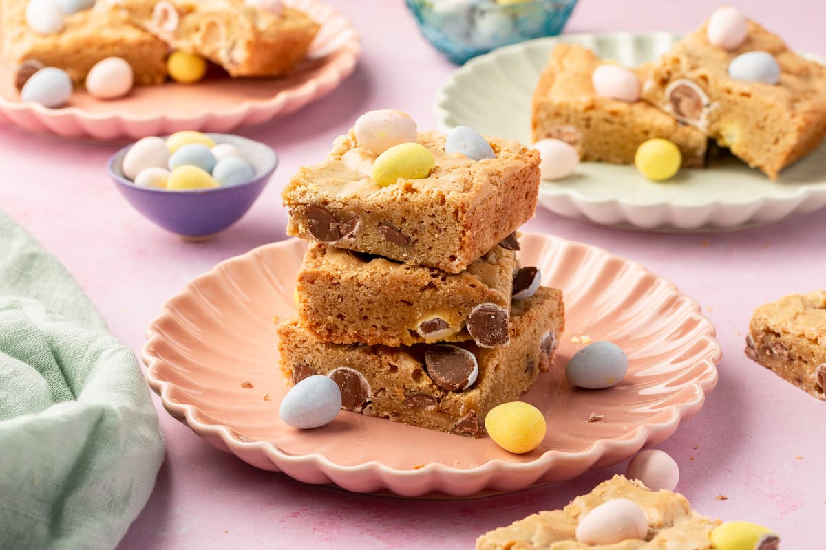 A stack of three gluten-free mini egg blondies on a pink scalloped plate with more blondies on plates in the background.
